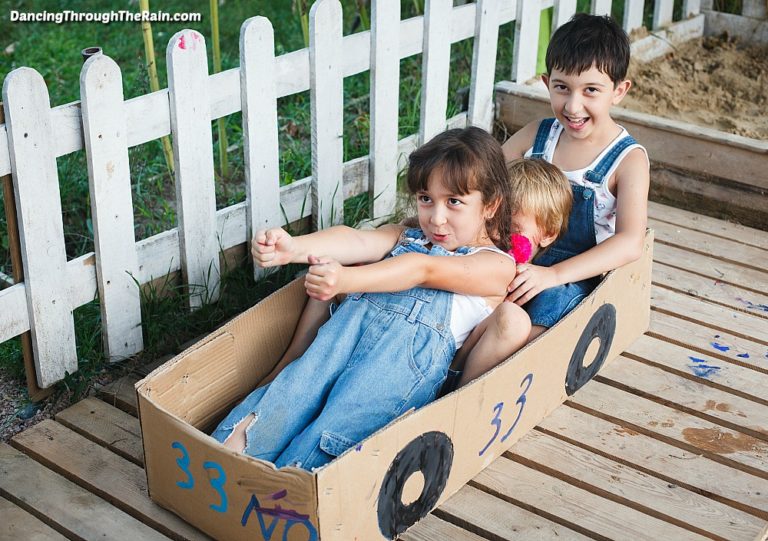 One picture of three children in a box that is drawn to look like a racecar on a wooden porch in front of a white picket fence