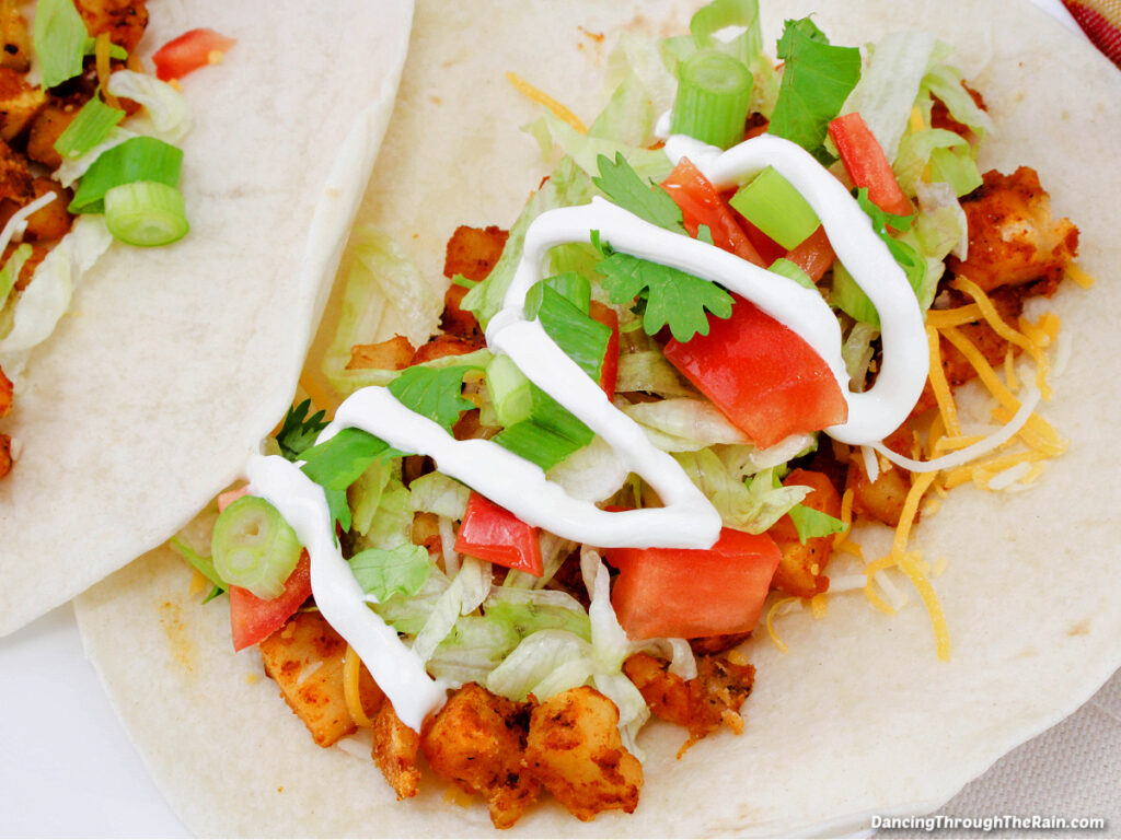 A closeup of a Crispy Potato Taco with visible sour cream, tomatoes, cilantro, shredded cheese, and seasoned potatoes