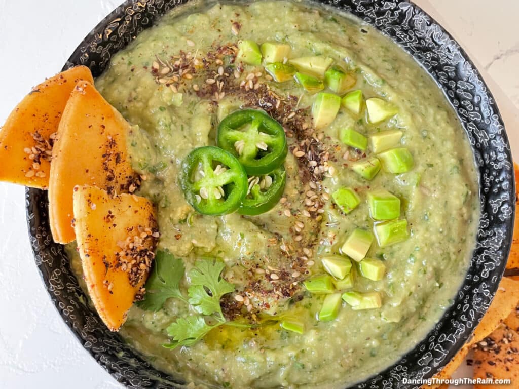 A black bowl with Avocado Cilantro Garlic Hummus with visible avocado pieces, sesame seeds, three jalapeno slices, and three pita chips inside