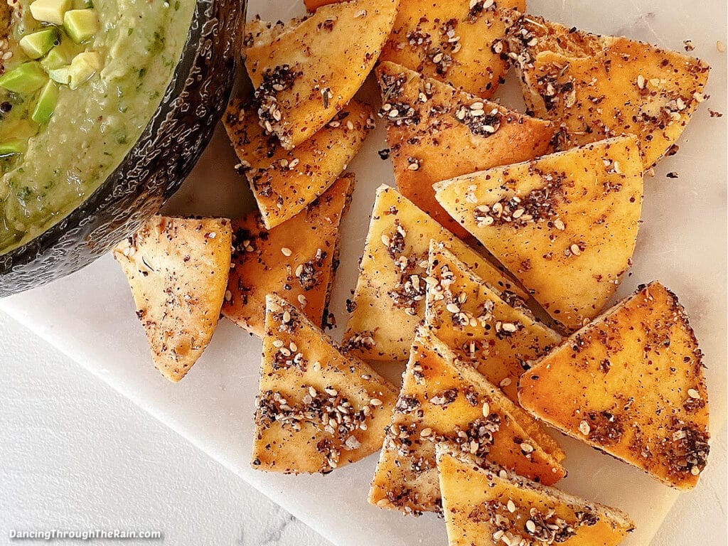 A closeup of Baked Za'atar Pita Chips on a white serving dish next to a black bowl with avocado hummus