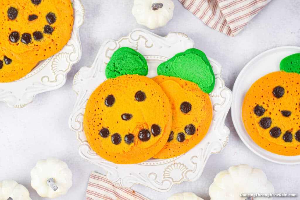 Four Jack-O-Lantern pancakes on white plates with small white pumpkins on a white table